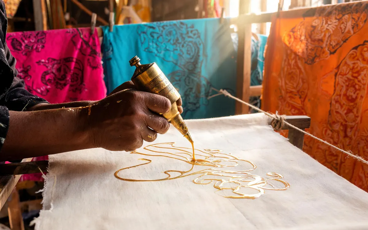 Foto hero laman web janaan AI: tukang batik melukis corak lilin di sebelah kain batik warna-warni